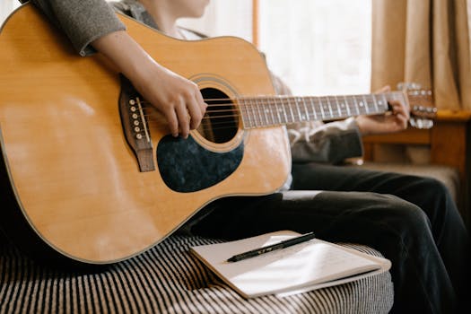 Person playing acoustic guitar with notebook and pen, reflecting songwriting process indoors.