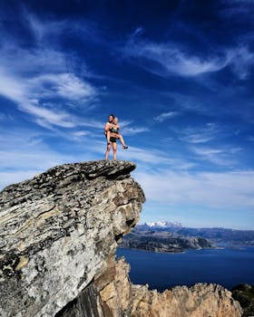 Two climbers atop a dramatic cliff with a stunning ocean view under a vibrant blue sky.