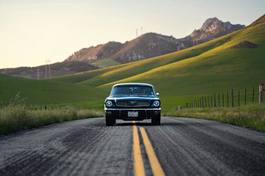 Old-fashioned car driving on a rural road with scenic green hills in the background.